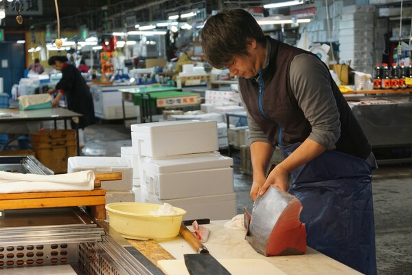 Young Japanese man preparing styrofoam boxes of fresh fish. Huge traditional knife on the table in front of him.